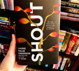 A woman's hand holding a copy of the book Shout by Laurie Halse Anderson
