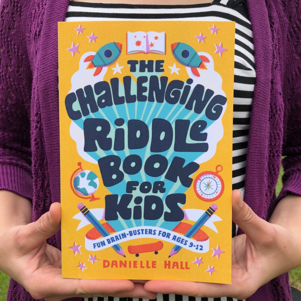 Danielle Hall is holding The Challenging Riddle Book for Kids which has a yellow cover and a joyful font.