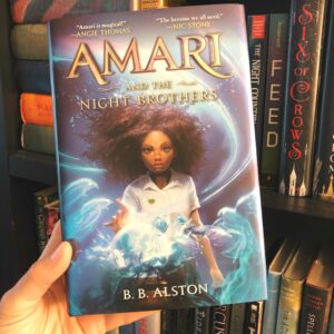 The book Amari and the Night Brothers by B.B. Alston held up in front of a book shelf.