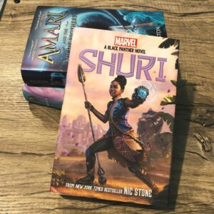 A bookstack on a woodgrain background, the book on top is Shuri by Nic Stone.